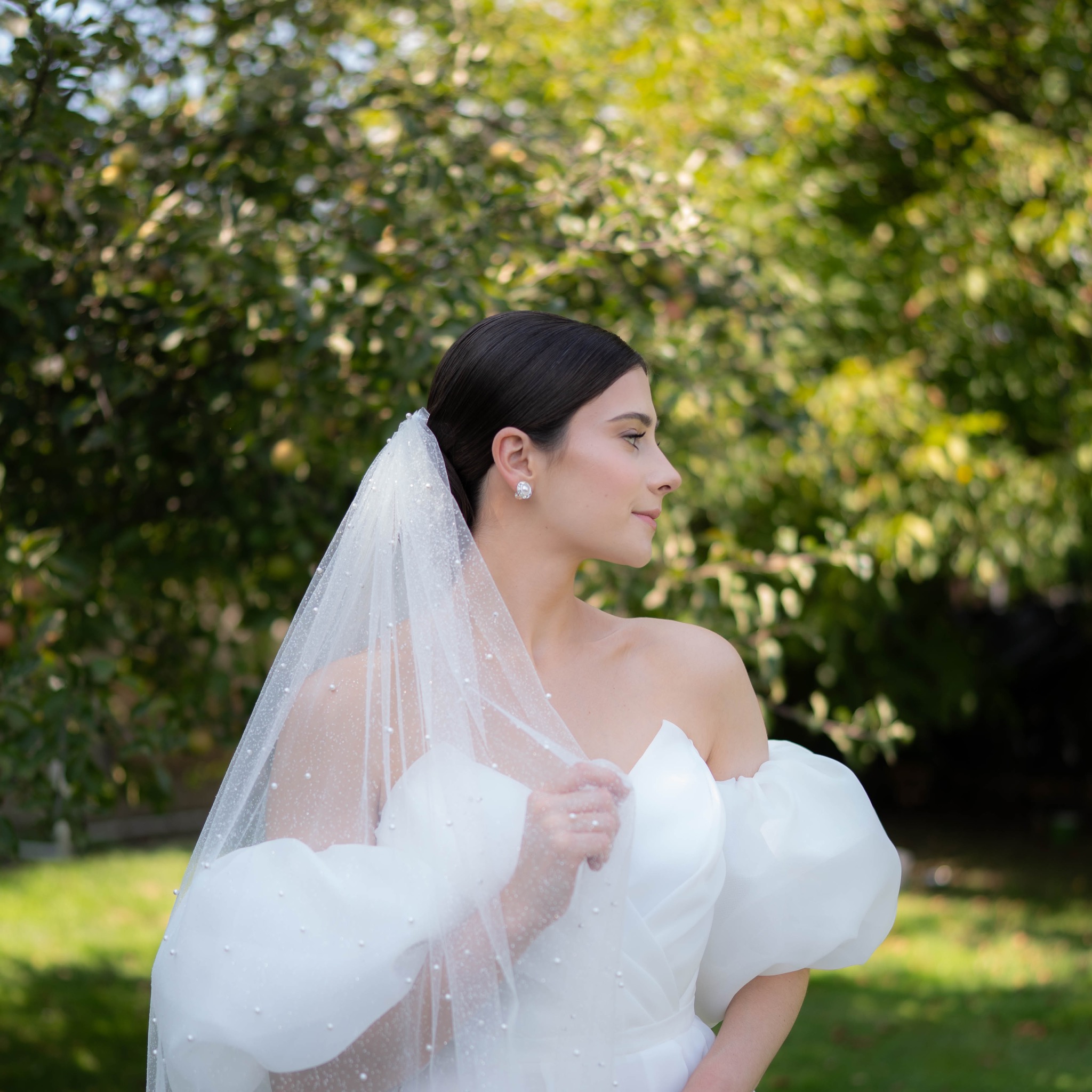 Elegancka panna młoda w białej sukni i welonie z perłami, z profilu, na tle zieleni. Subtelne oświetlenie podkreśla detale stroju i naturalne piękno.