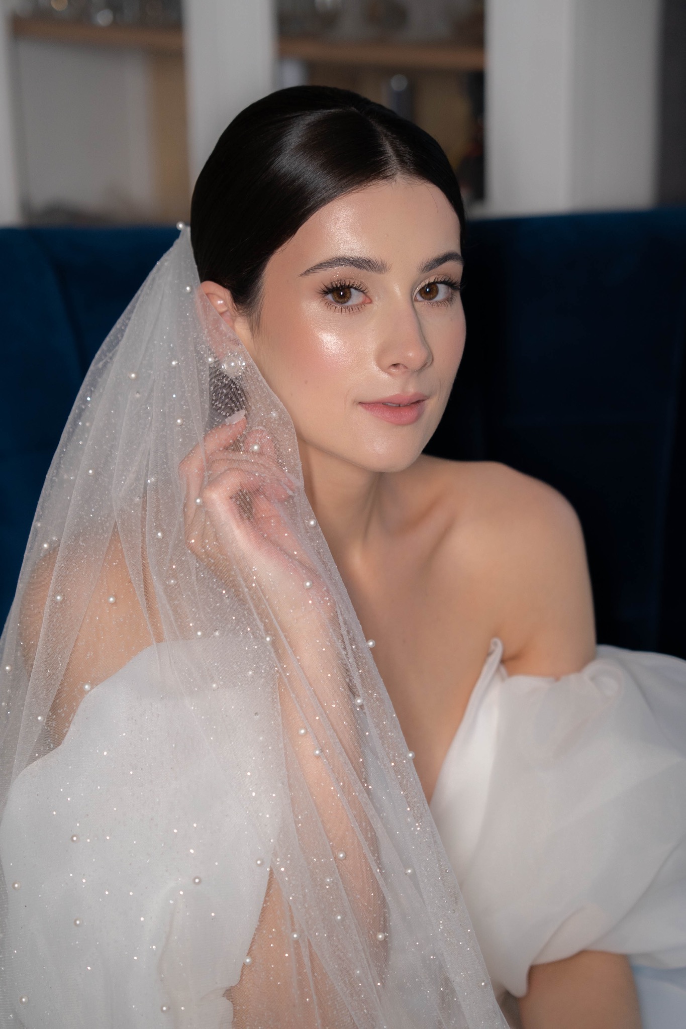 Portret panny młodej w welonie z perłami, delikatny makijaż, gładko upięte włosy, na tle ciemnego fotela. Subtelne ujęcie podkreśla elegancję i blask.