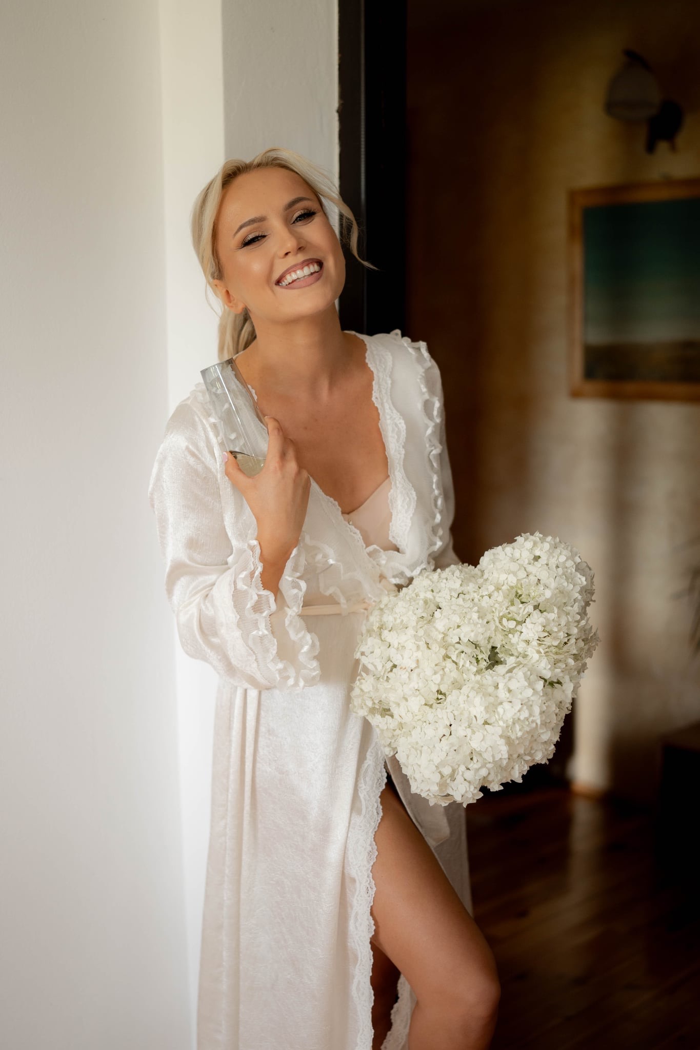 Uśmiechnięta blondynka w szlafroku z bukietem białych kwiatów i kieliszkiem w dłoni, oparta o białą ścianę, z widocznym fragmentem wnętrza w tle.