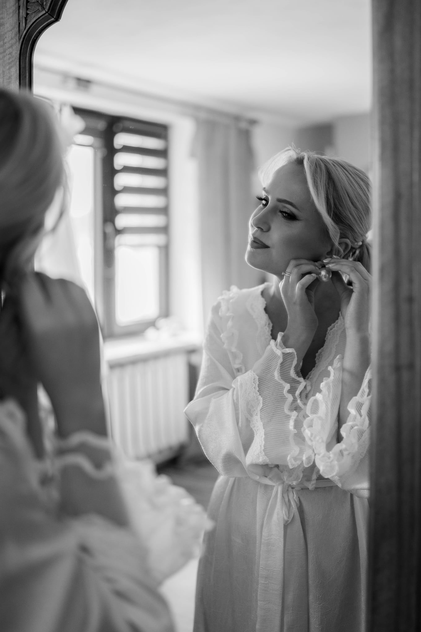 Czarno-białe zdjęcie kobiety w szlafroku, zakładającej kolczyk przed lustrem. Widoczne okno z żaluzjami w tle. Delikatne światło podkreśla detale.