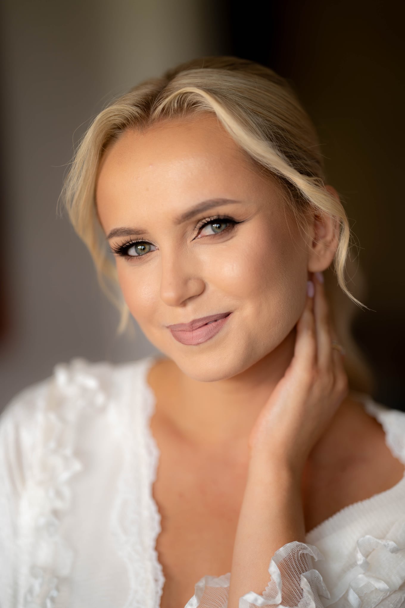 Portret kobiety o blond włosach i zielonych oczach w białym szlafroku z koronką, delikatny makijaż, dłoń przy szyi, subtelny uśmiech, miękkie światło.