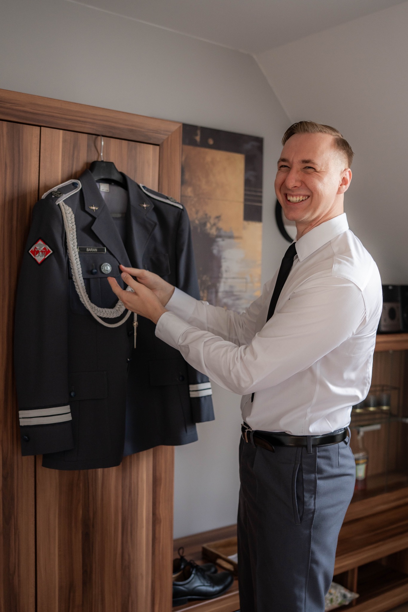 Uśmiechnięty mężczyzna w koszuli i spodniach, obok munduru galowego wiszącego na szafie. Przygotowania do uroczystości.