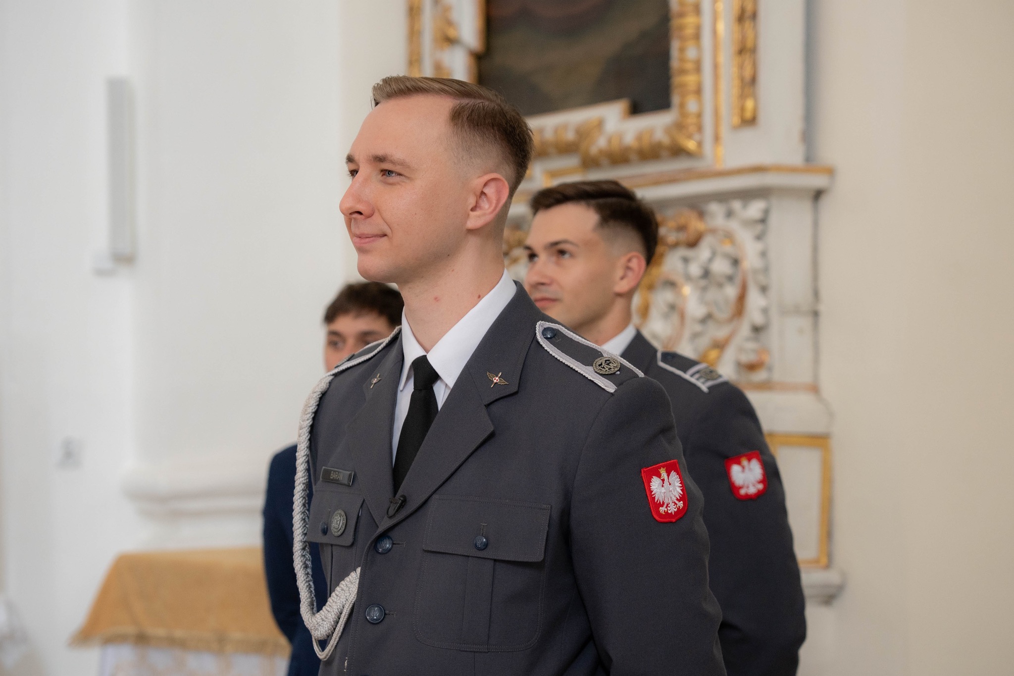 Portret młodego mężczyzny w mundurze galowym Wojska Polskiego, z naszywką z godłem, na tle jasnego wnętrza kościoła z dekoracjami.
