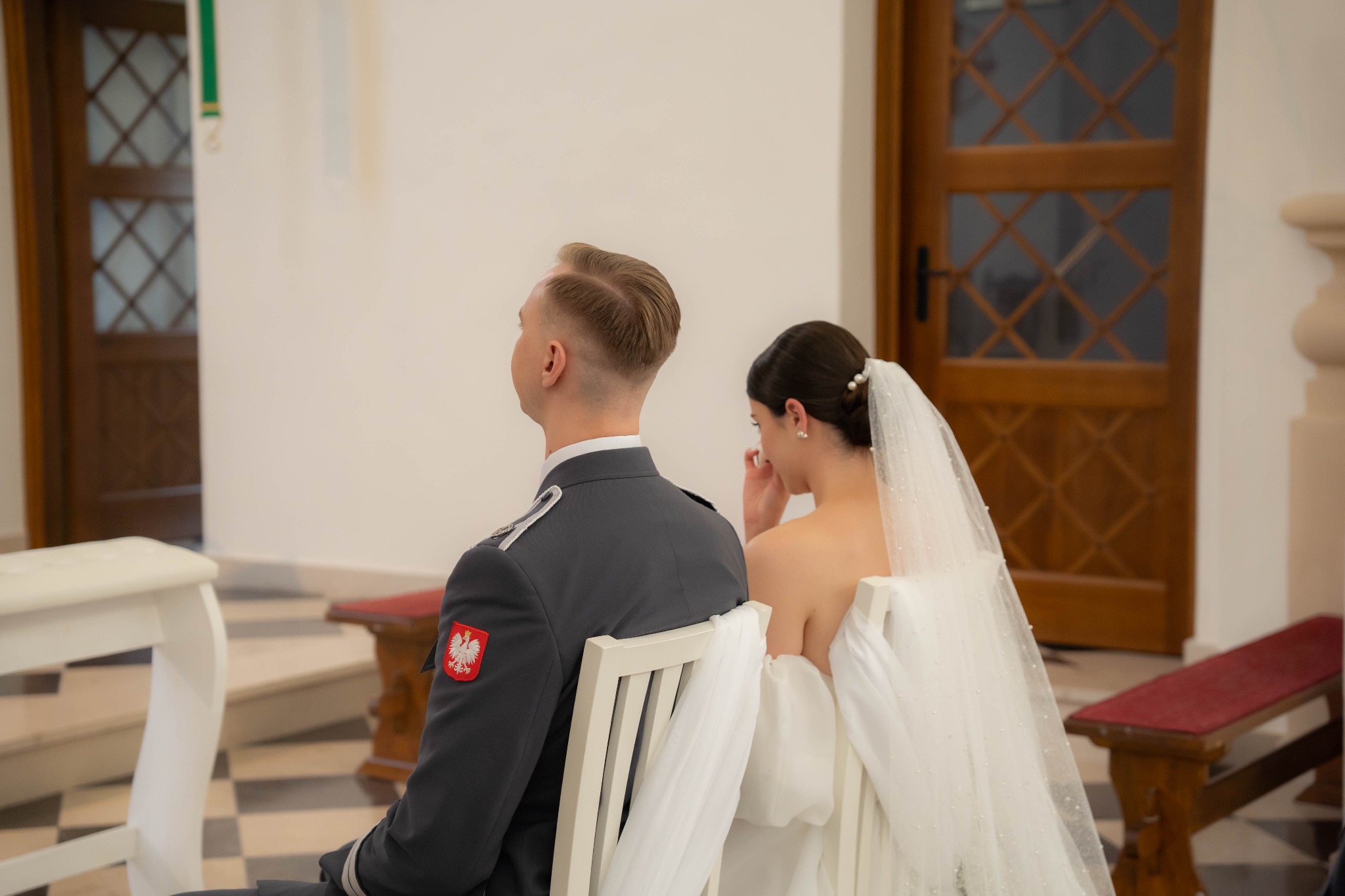 Para młoda siedzi w kościele: pan młody w mundurze Wojska Polskiego, panna młoda w białej sukni i welonie. Ujęcie z tyłu, widoczna łza wzruszenia panny młodej.