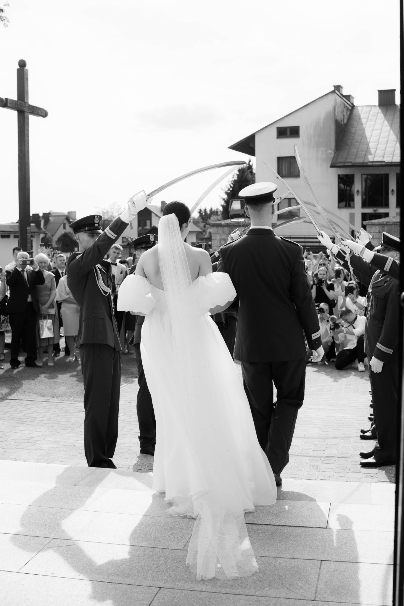 Czarno-białe zdjęcie zza pleców panny młodej w sukni ślubnej, przechodzącej pod szpalerem szabel trzymanych przez żołnierzy w mundurach. W tle budynek i obserwujący ludzie.