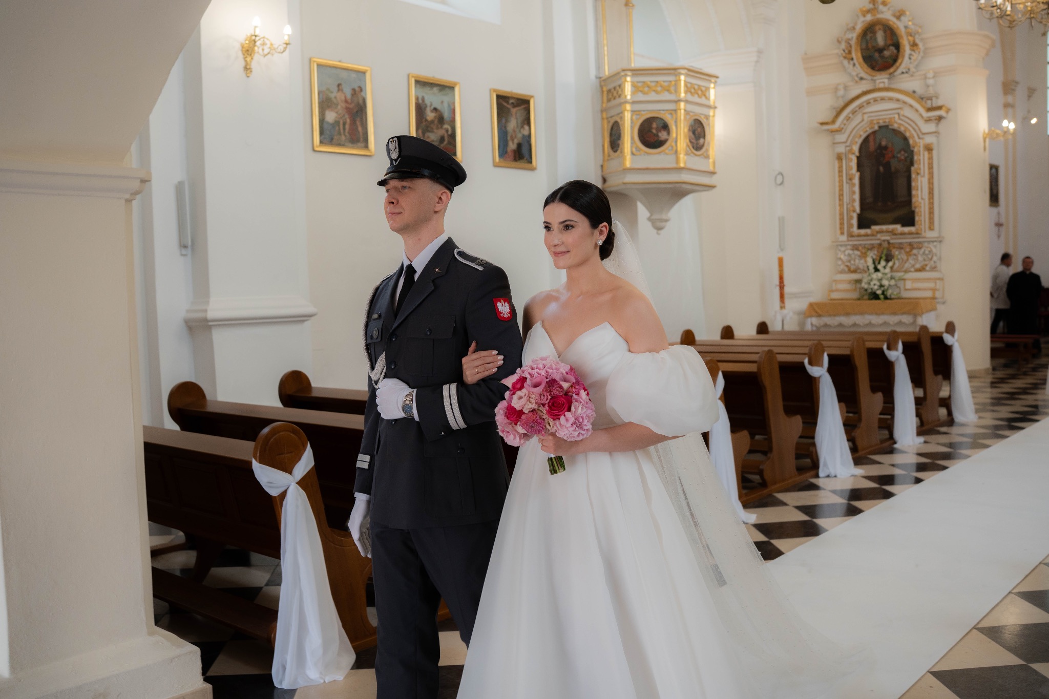 Panna młoda z bukietem i mężczyzna w mundurze galowym idą w kościele. Uwagę przykuwa dekoracja ławek białymi wstążkami i dywan w czarno-białą szachownicę.