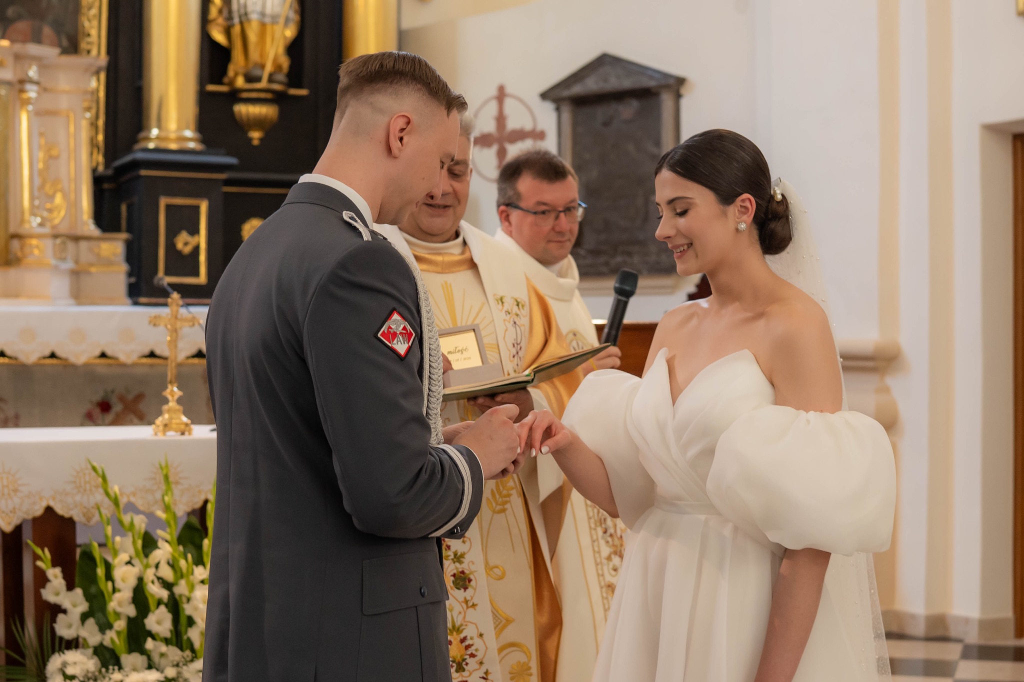 Moment zakładania obrączki podczas ceremonii ślubnej w kościele. Panna młoda w białej sukni, pan młody w mundurze. W tle ksiądz z mikrofonem i otwartą księgą.