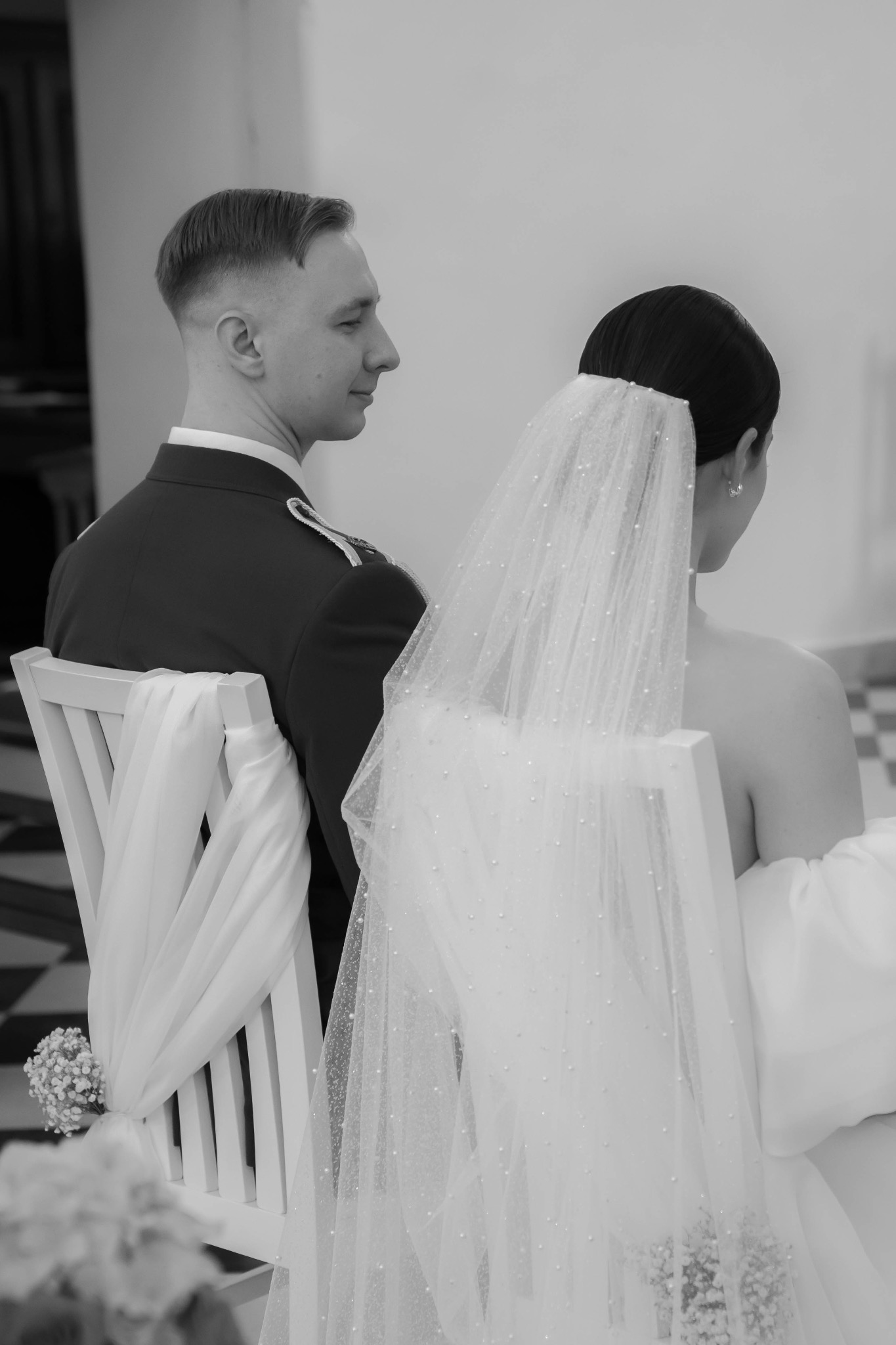 Czarno-białe zdjęcie pary młodej w kościele, siedzącej na krzesłach z dekoracjami, widok z tyłu, panna młoda w welonie z perełkami, pan młody w mundurze.