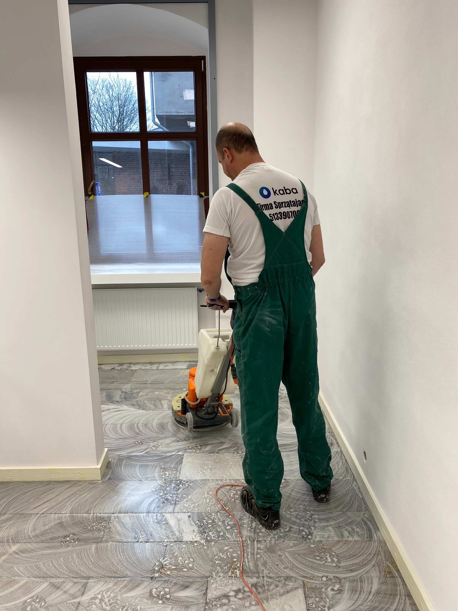 Pracownik w zielonym kombinezonie czyści podłogę z płytek maszyną czyszczącą w jasnym pomieszczeniu. Widoczny napis na koszulce firmy sprzątającej.
