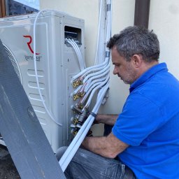 Feniks Kędra Krzysztof - Montaż klimatyzacji zewnętrznej Rotenso przez instalatora w niebieskiej koszulce. Widoczne rury i złącza. Budynek w tle.