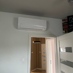 Feniks Kędra Krzysztof - Biały klimatyzator Rotenso Revio zamontowany na ścianie w jasnym pokoju, widok z perspektywy drzwi do innego pomieszczenia z tapetą w paski i lampą.