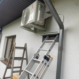 Feniks Kędra Krzysztof - Montaż klimatyzatora Rotenso na elewacji domu w Łukowie. Widoczne dwie drabiny, rura spustowa i okno. Biała elewacja z fakturą baranka.