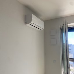 Feniks Kędra Krzysztof - Biały klimatyzator marki Tensa zamontowany na ścianie w jasnym pomieszczeniu z otwartym oknem i widokiem na budynek. Minimalistyczny design, nowoczesne wnętrze.
