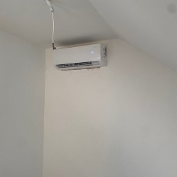 Feniks Kędra Krzysztof - Biała jednostka klimatyzacyjna zamontowana na ścianie w jasnym pomieszczeniu, widoczne przewody podłączeniowe. Minimalistyczny design, nowoczesne rozwiązanie.