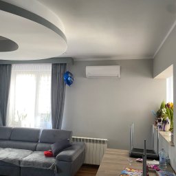 Feniks Kędra Krzysztof - Biała klimatyzacja ścienna w szarym salonie z sofą, zasłonami i dekoracyjnym sufitem. Na ścianie wisi niebieski balon. Widoczny grzejnik i stół z przedmiotami.