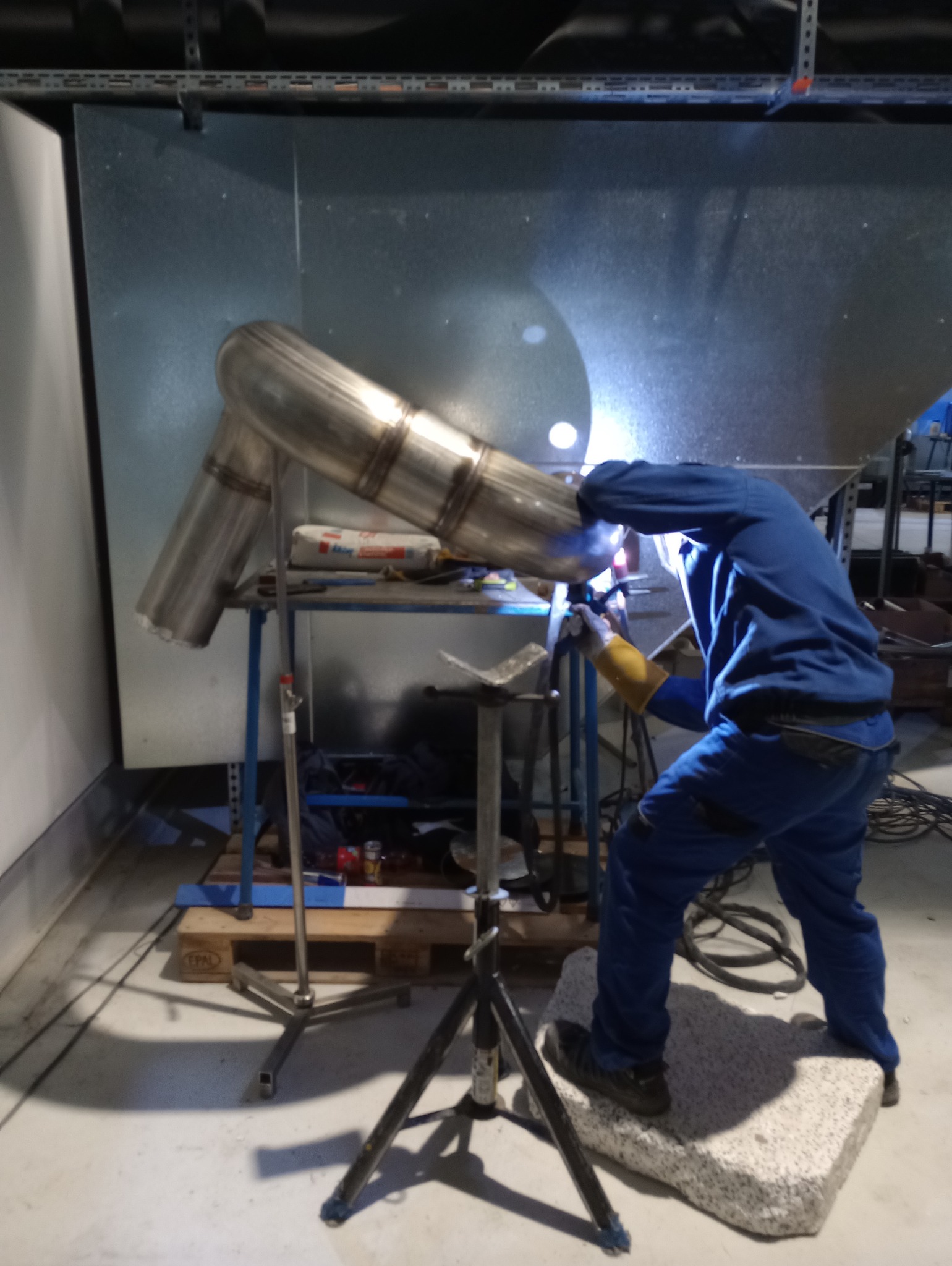 Spawacz w niebieskim kombinezonie łączy elementy rury ze stali nierdzewnej w warsztacie, używając spawarki. Widoczne iskry i wyposażenie spawalnicze.
