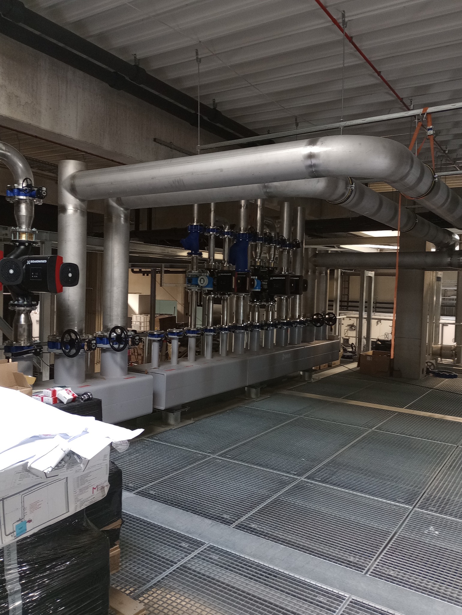 Instalacja przemysłowa: srebrne rury, zawory i pompy w hali. Widoczna metalowa kratownica podłogowa i dokumentacja techniczna. Elementy spawane.