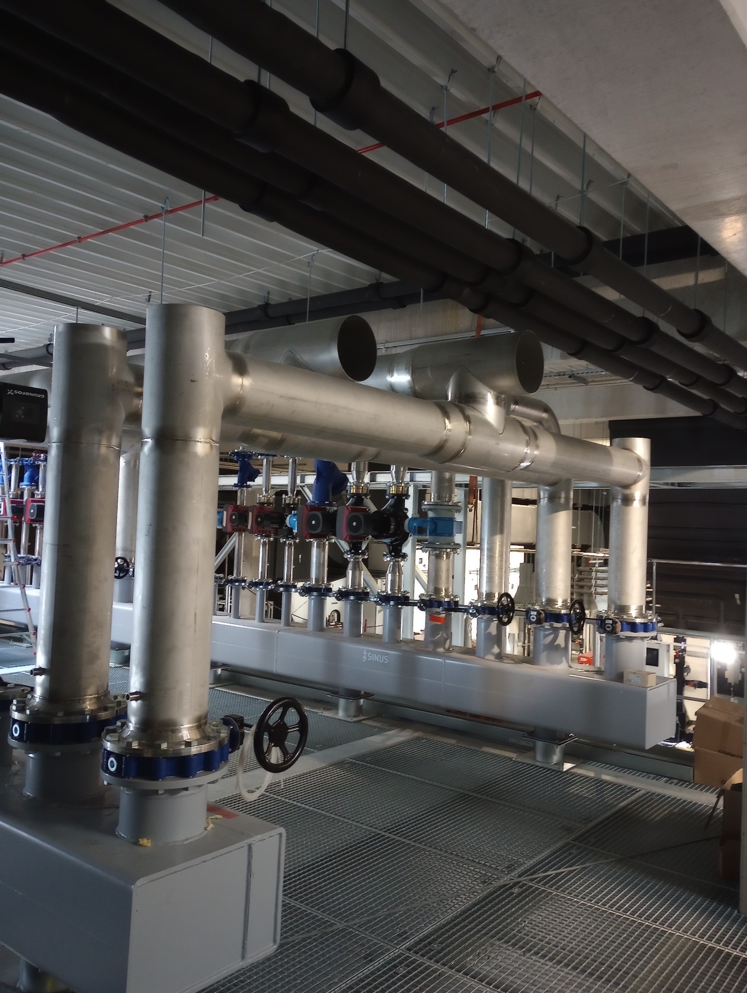 Kompleksowa instalacja przemysłowa z rurami, zaworami i pompami, z metalową kratą podłogową i czarnymi rurami pod sufitem. Elementy instalacji ze stali nierdzewnej.