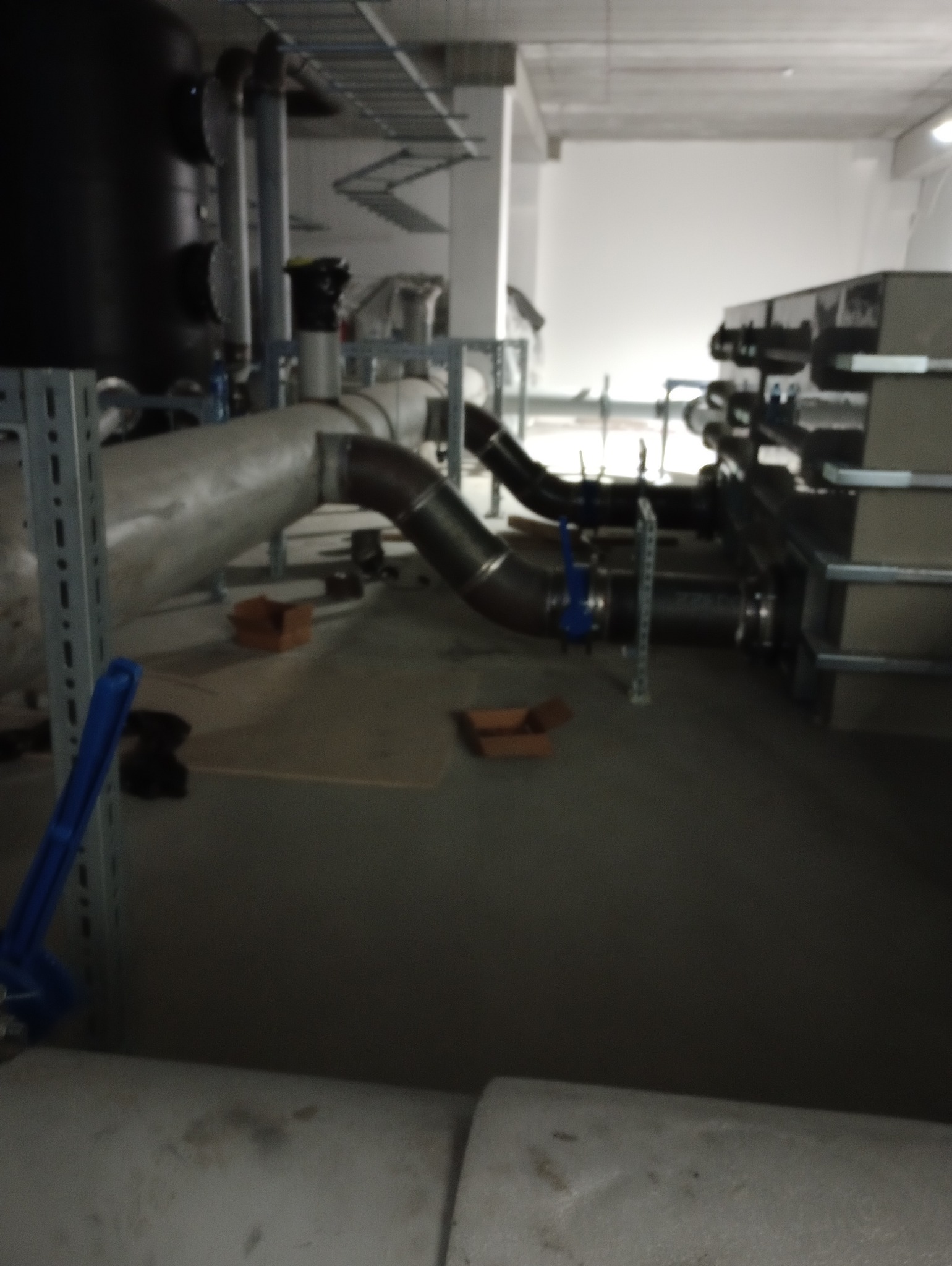 Instalacja przemysłowa: rury ze złączkami, zawory i podwieszane koryta kablowe w hali. Widoczne podpory i elementy mocujące.