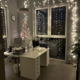 Klimatyczne biuro z dekoracją świetlną: biurko z komputerem, krzesło, zegar na ścianie i okno z widokiem na miasto, ozdobione lampkami. Świąteczny nastrój w miejscu pracy.