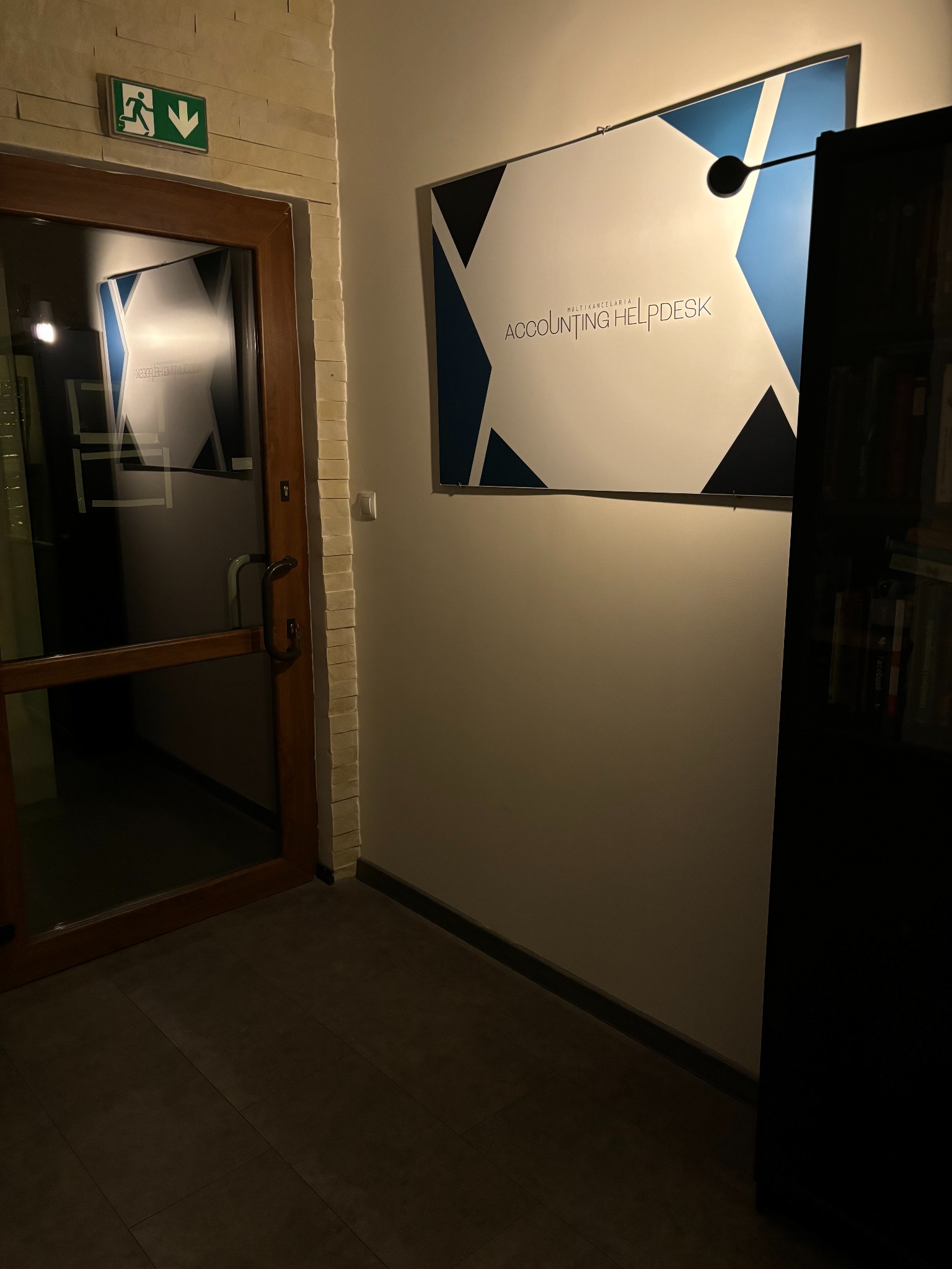 Wnętrze biura z plakatem 'Accounting Helpdesk' na ścianie, widoczne drzwi z piktogramem wyjścia ewakuacyjnego i fragment regału z książkami. Stonowana kolorystyka.