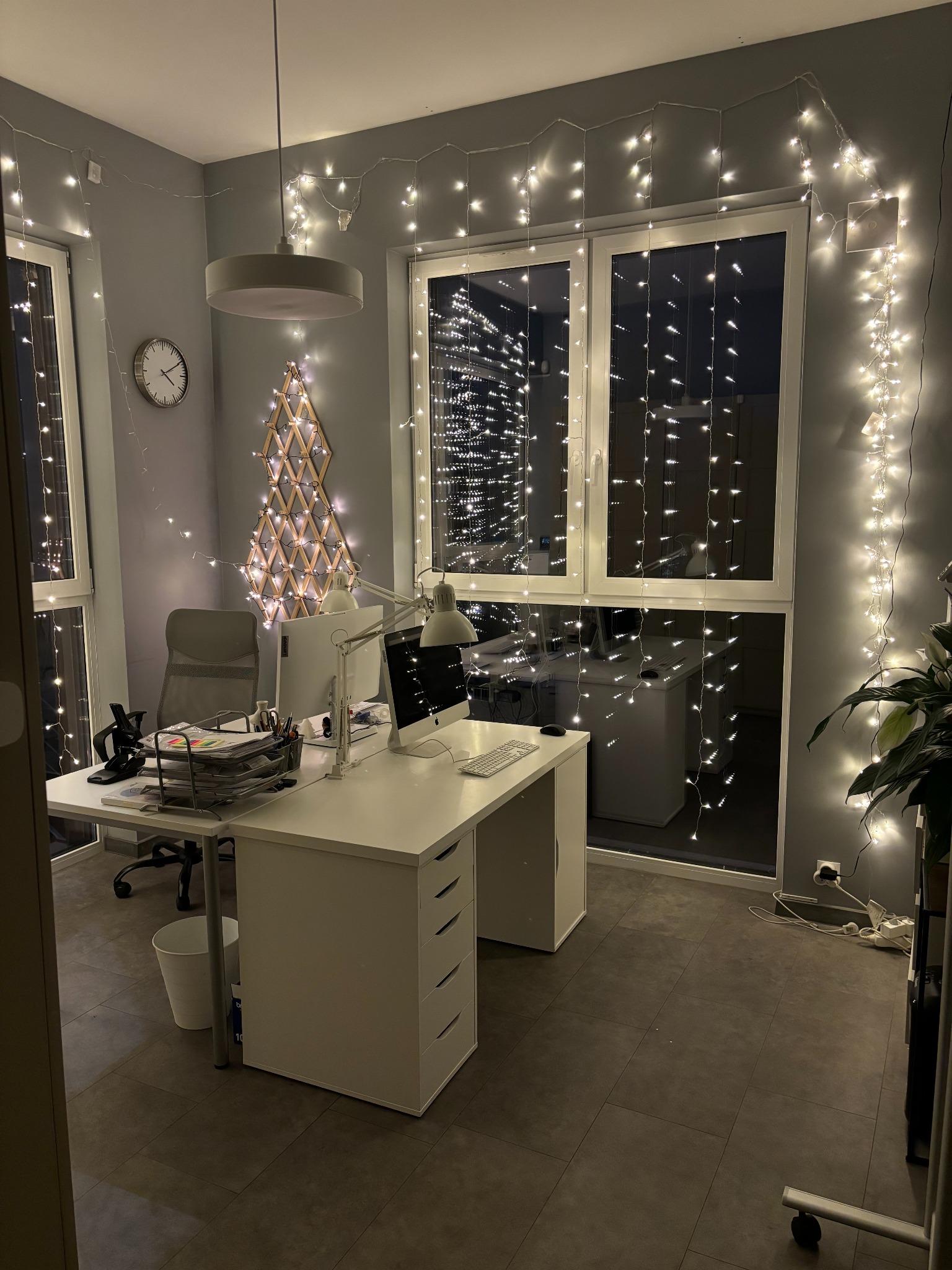 Klimatyczne biuro z dekoracją świetlną: biurko z komputerem, krzesło, zegar na ścianie i okno z widokiem na miasto, ozdobione lampkami. Świąteczny nastrój w miejscu pracy.