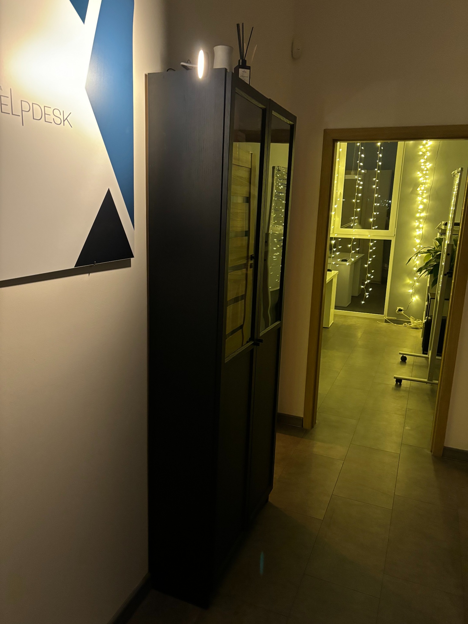 Nowoczesne biuro: minimalistyczny design z grafiką 'Helpdesk', czarną szafą i widokiem na pokój z dekoracyjnymi lampkami przez otwarte drzwi.