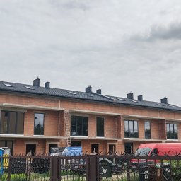 DOMI-BUD - Budowa szeregowca z czerwonej cegły, widoczne okna, drabina oparta o budynek, częściowo zadaszony dach, w tle pochmurne niebo. Etap murowania ścian.