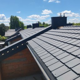 DomiBud Dominik Pośpiech - Szary dach z dachówki ceramicznej w trakcie budowy, widoczne kominy i fragmenty murowanych ścian. Błękitne niebo w tle. Dachówka ułożona równo.