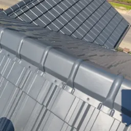 Szary dach z blachy trapezowej po montażu, widoczne łączenia i mocowania. W tle fragment dachu z dachówki ceramicznej.