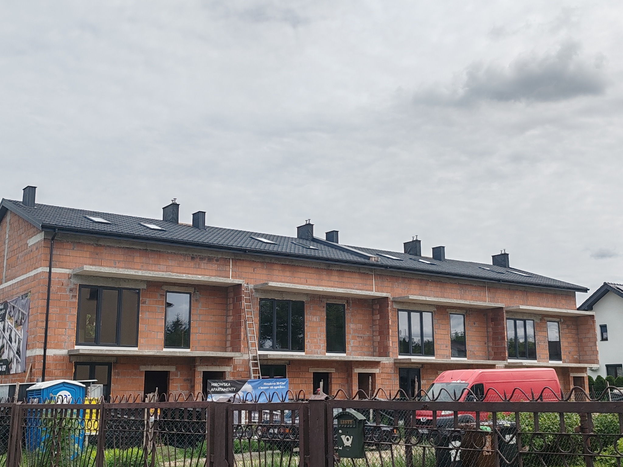 Budowa szeregowca z czerwonej cegły, widoczne okna, drabina oparta o budynek, częściowo zadaszony dach, w tle pochmurne niebo. Etap murowania ścian.