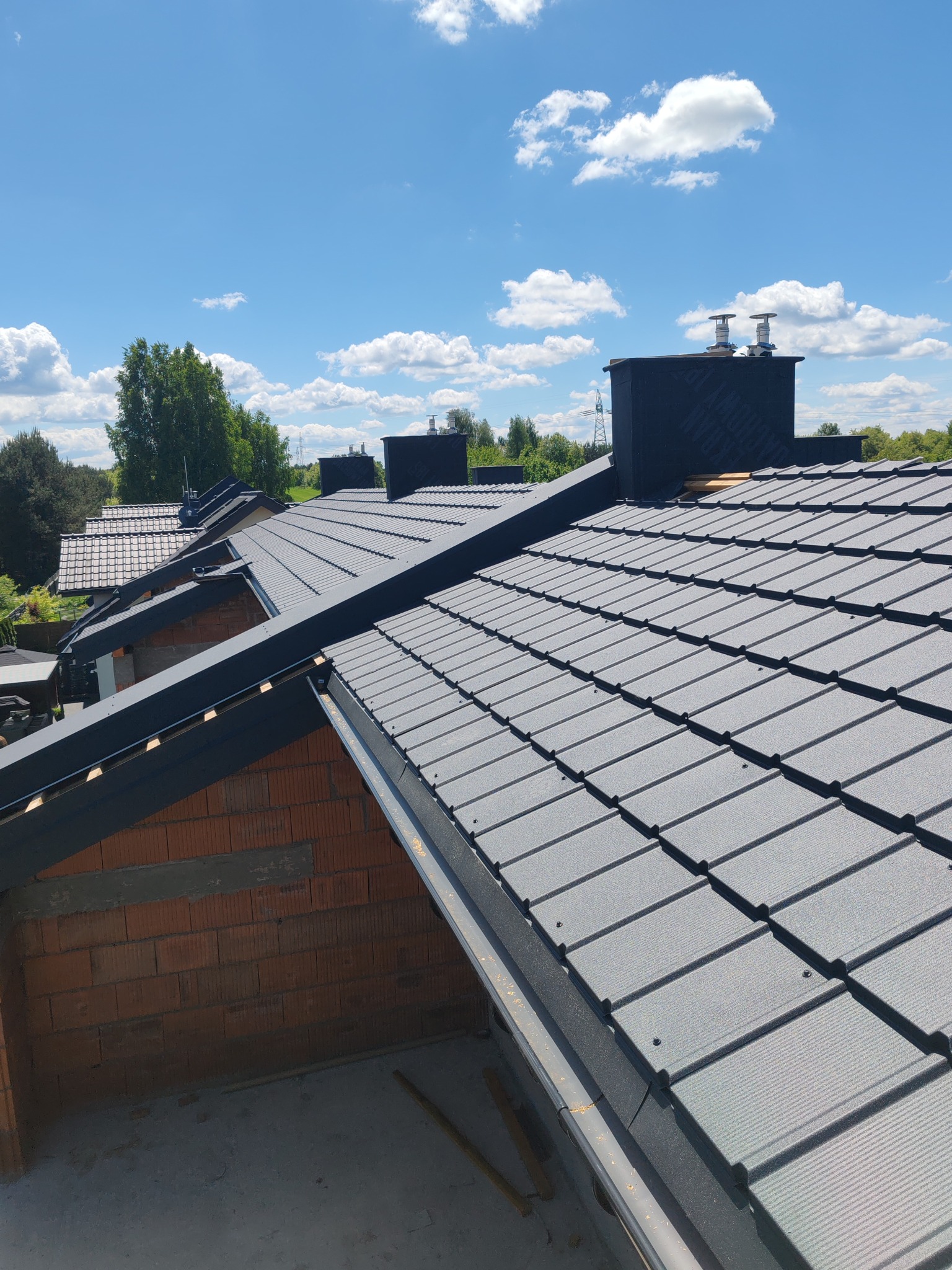 Szary dach z dachówki ceramicznej w trakcie budowy, widoczne kominy i fragmenty murowanych ścian. Błękitne niebo w tle. Dachówka ułożona równo.