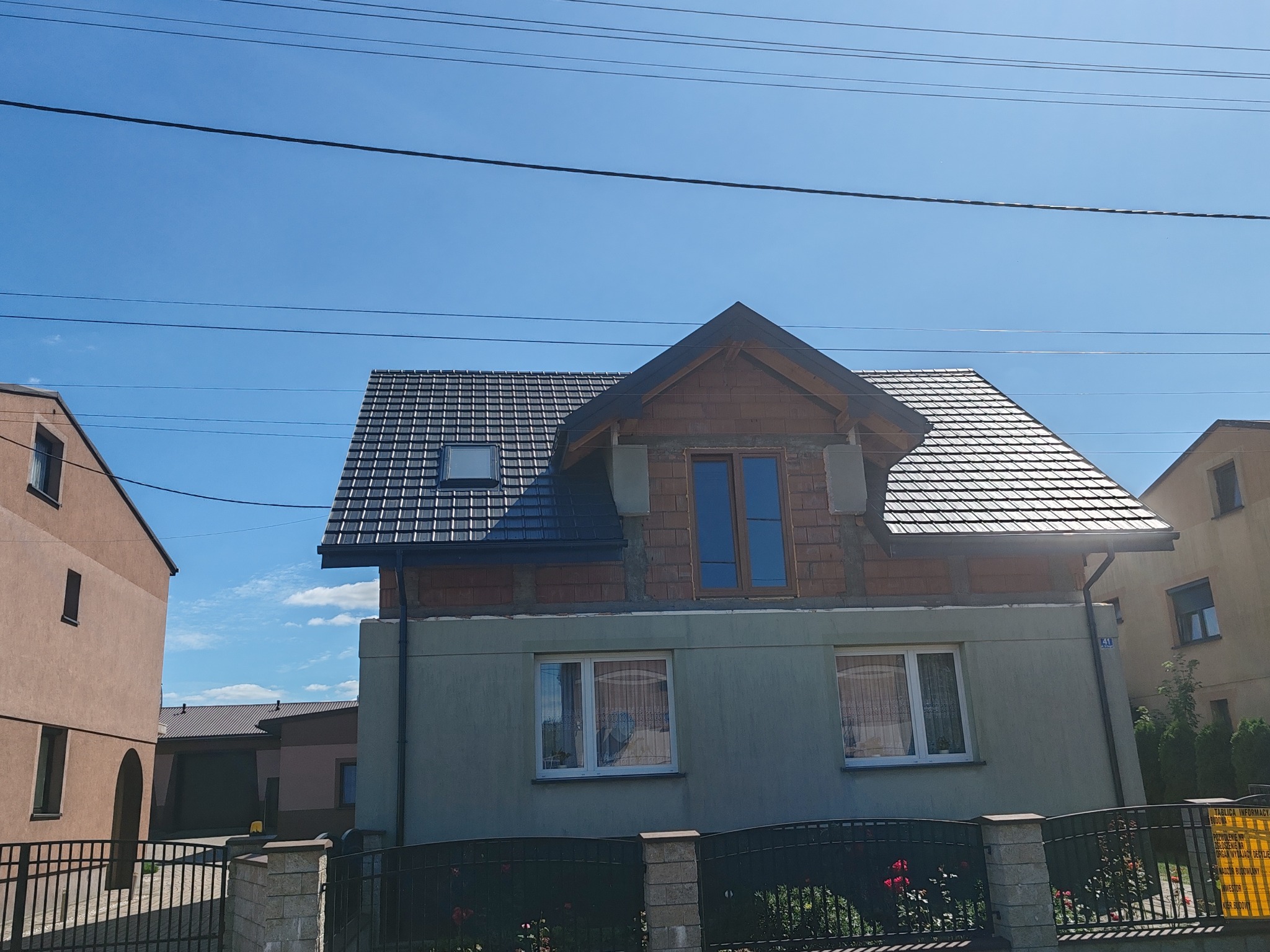 Dom w trakcie budowy: ciemna dachówka, ceglana ściana na parterze z oknami, błękitne niebo w tle. Widoczne przewody elektryczne.