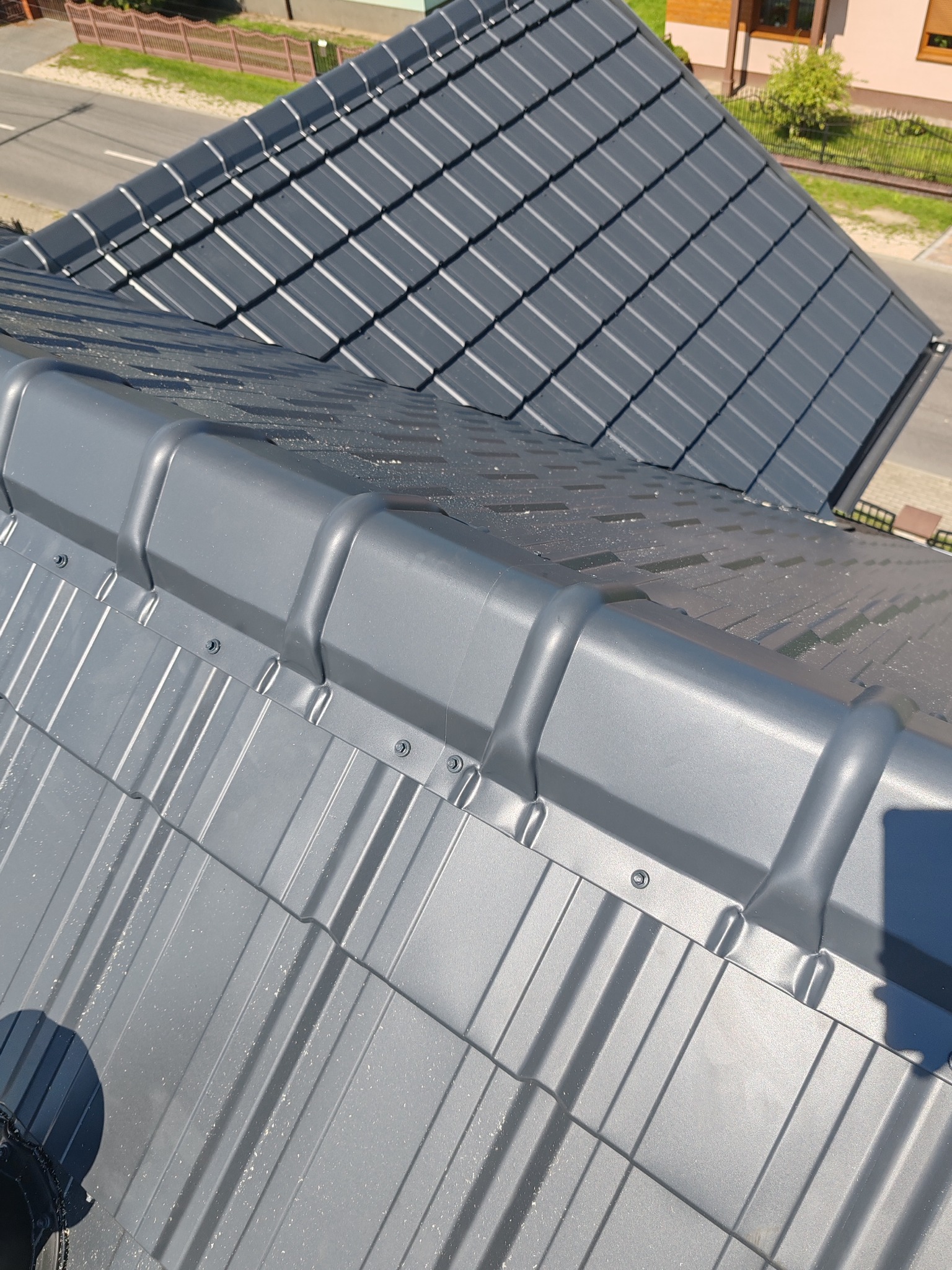 Szary dach z blachy trapezowej po montażu, widoczne łączenia i mocowania. W tle fragment dachu z dachówki ceramicznej.