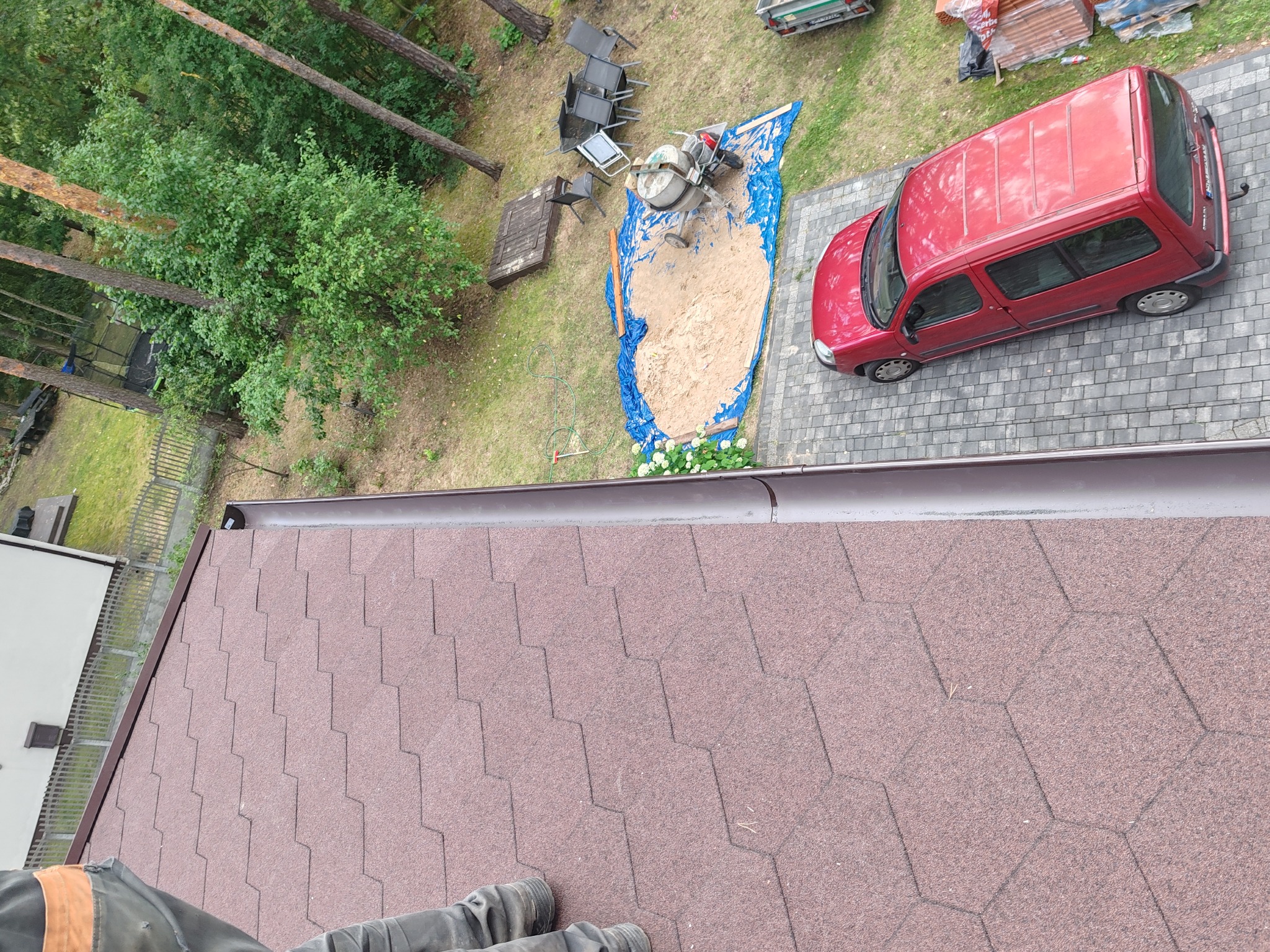 Widok z góry na nowo położony dach z bordowej gontu bitumicznego, z widocznymi fragmentami rynny i sylwetką osoby w dolnej części kadru. W tle zieleń, samochód i betoniarka.
