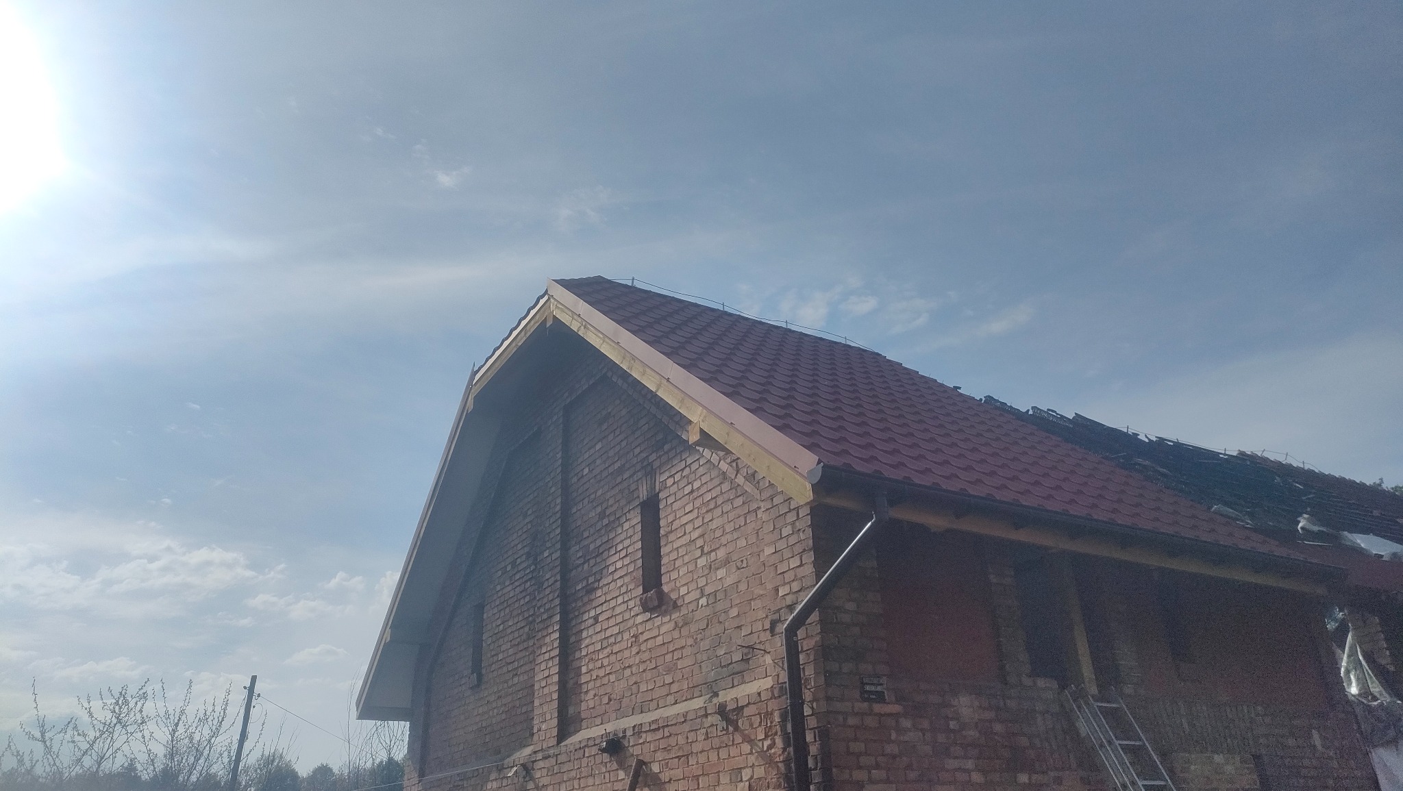 Częściowo pokryty dach z czerwonej dachówki na murowanym budynku z cegły, widoczne krokwie, fragment starego pokrycia. Błękitne niebo w tle, słoneczny dzień.