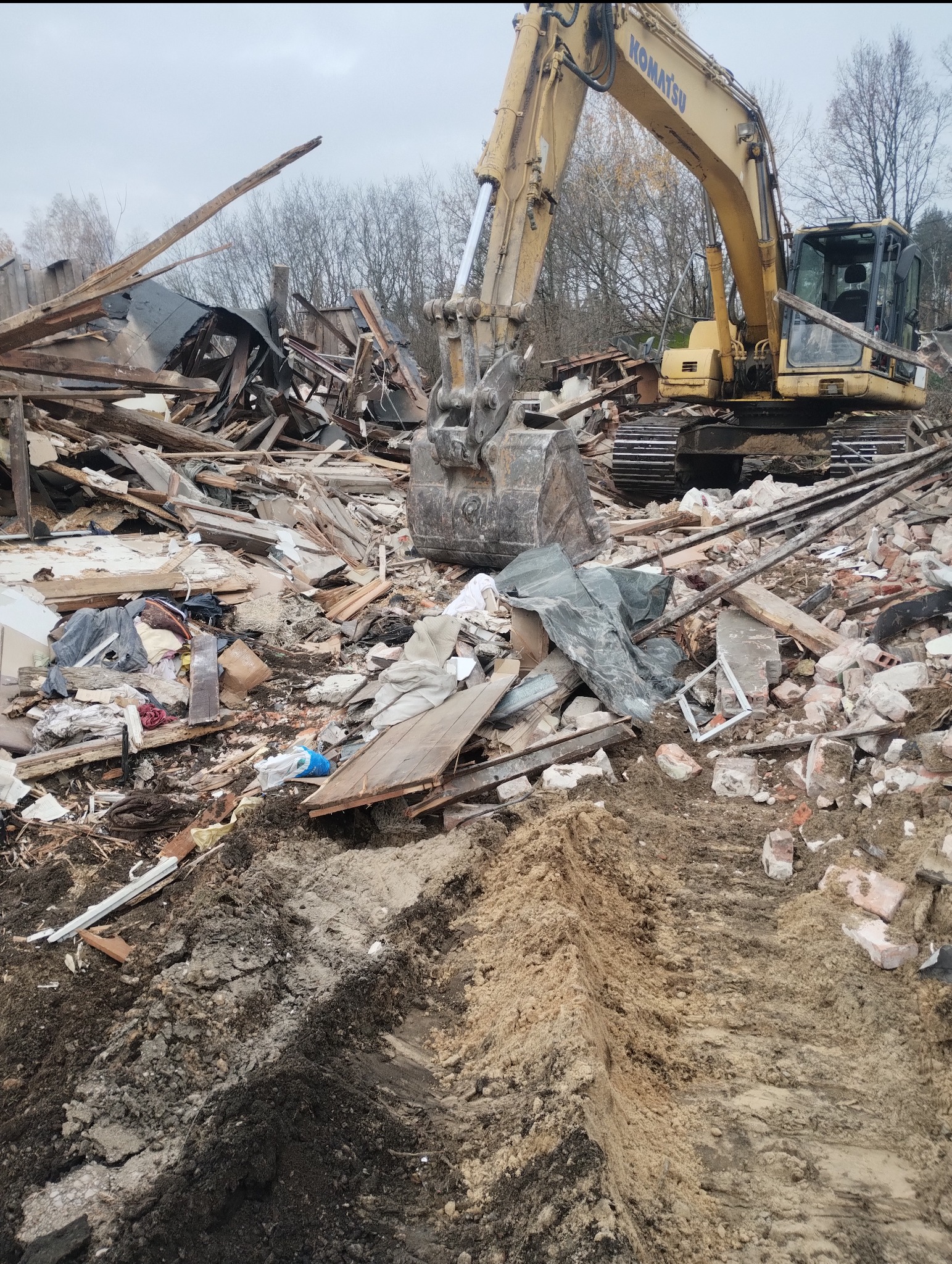 Żółta koparka Komatsu na gruzowisku po rozbiórce budynku w Legionowie. Widoczne resztki konstrukcji i ślady opon na ziemi.