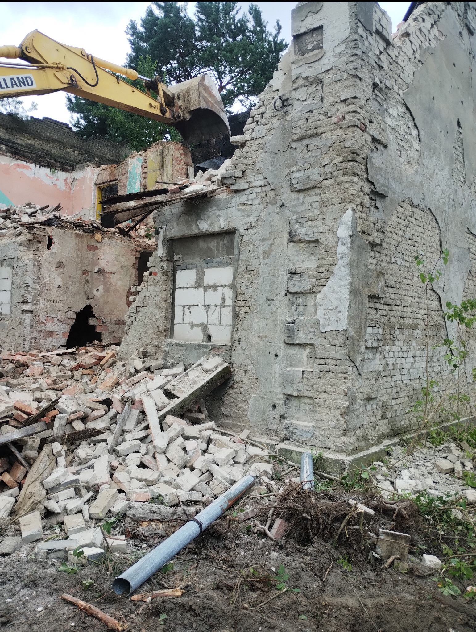 Żółta koparka z łyżką w trakcie rozbiórki starego, ceglanego budynku. Widoczne gruzy i fragmenty ścian. Ujęcie z dołu, zieleń w tle.