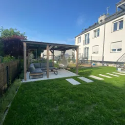 Nowoczesna altana z szarą sofą i wiszącym fotelem na zadbanym trawniku w ogrodzie miejskim, z widokiem na budynek mieszkalny w tle.