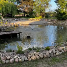 Oczko wodne z fontanną i drewnianym pomostem, otoczone kamieniami i zielenią. Uroku dodają drzewa i rośliny w tle, tworząc sielski krajobraz.