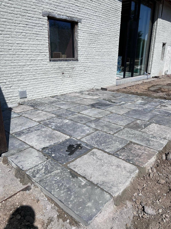 Ułożona kostka brukowa z szarego kamienia na tarasie przy białym, ceglanym budynku. Widoczny fragment ogrodu i okno z drewnianą ramą.