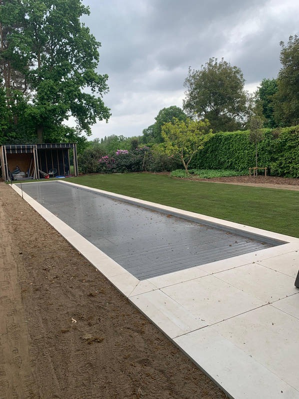 Nowoczesny ogród z równo przystrzyżonym trawnikiem i ukrytą podziemną instalacją. Widoczna jasna kostka brukowa i budowa w tle.