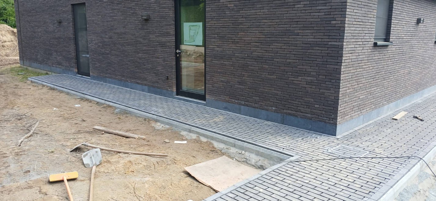 Ułożona kostka brukowa wzdłuż budynku z ciemnej cegły. Widoczne narzędzia brukarskie i fragmenty niezakończonych prac ziemnych. Kątowe ujęcie.