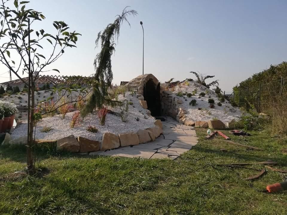 Ogród z kamienną grotą, białym żwirem i ścieżką z płyt. W tle widoczne drzewa i krzewy, oświetlone słońcem. Elementy dekoracyjne, rośliny ozdobne i trawa.