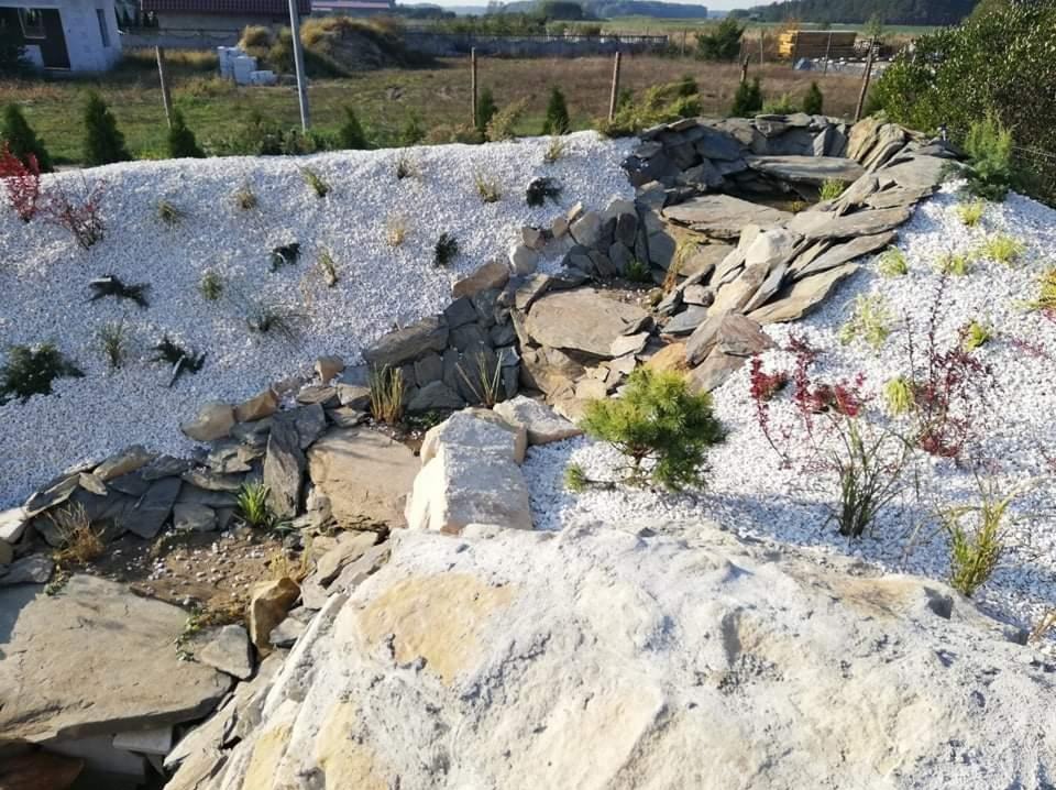 Ogród z kaskadą wodną wyłożoną kamieniami i białym grysem. W tle widoczne budynki i zieleń. Ujęcie z bliska, podkreślające detale aranżacji.