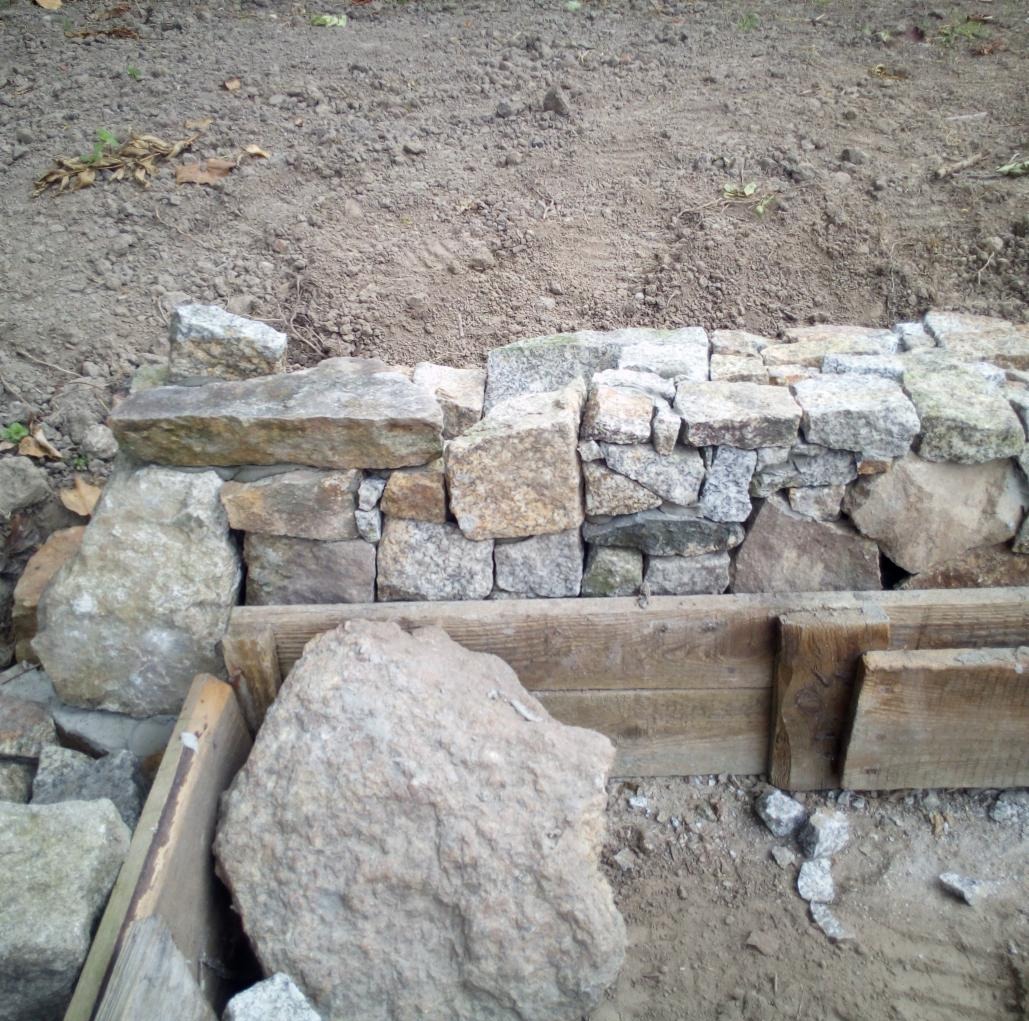 Budowa murku z kamienia polnego, widoczne szalunki drewniane. Częściowo ułożone kamienie na tle ziemi.