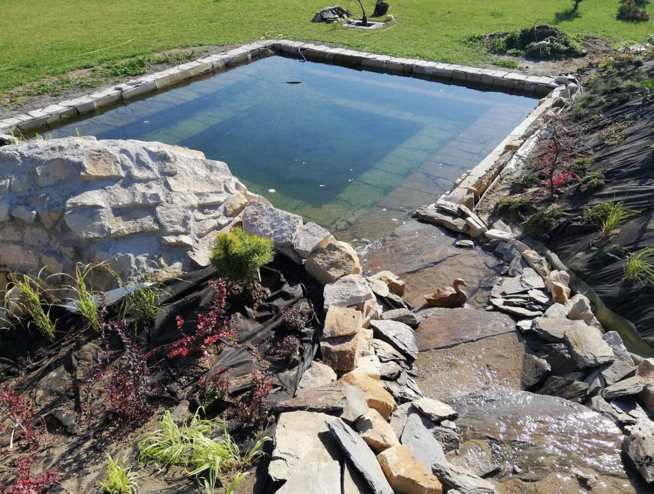 Oczko wodne z kamienną obudową i kaskadą wodną, otoczone roślinnością. Woda w zbiorniku jest czysta, a na dnie widać stopnie. Dekoracyjna kaczka w strumieniu.