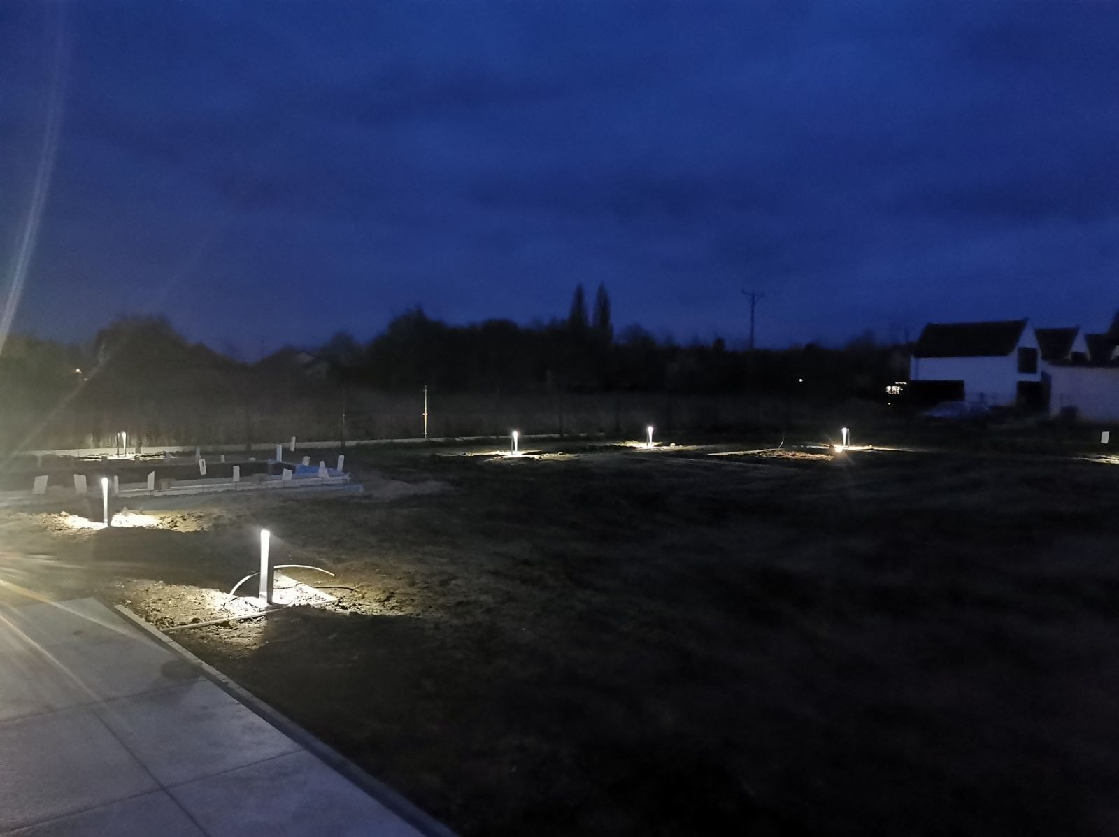 Nocny widok na teren z fundamentami i oświetleniem ogrodowym. Słupki świetlne równomiernie rozmieszczone, tworzą iluminację. W tle domy i ciemne niebo.