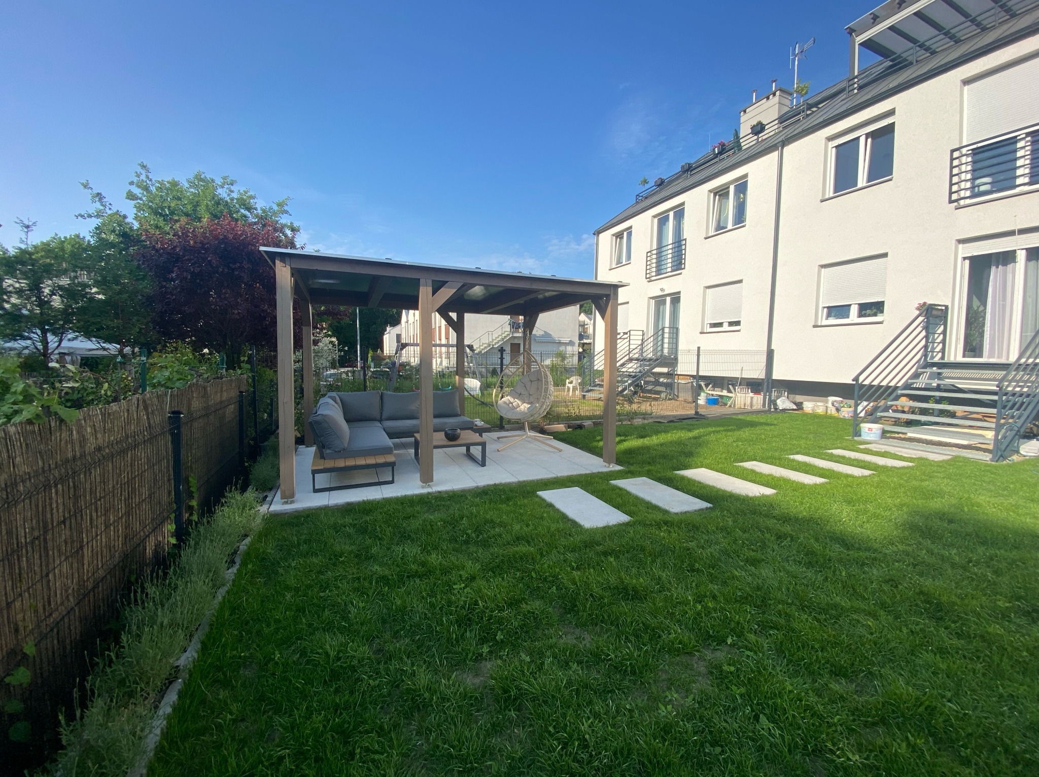 Nowoczesna altana z szarą sofą i wiszącym fotelem na zadbanym trawniku w ogrodzie miejskim, z widokiem na budynek mieszkalny w tle.