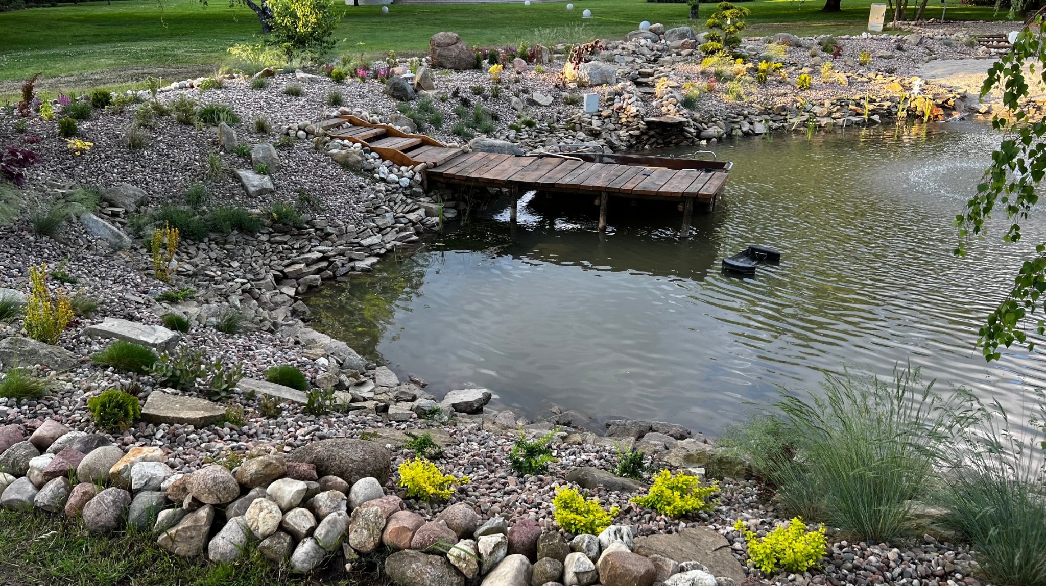 Oczko wodne z drewnianym pomostem, otoczone kamieniami i roślinnością. Woda odbija światło, tworząc spokojną atmosferę. Elementy małej architektury ogrodowej.